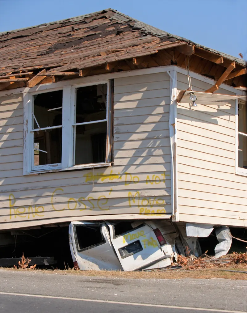 A wood-frame house knocked off its foundation by an earthquake, resting on a crushed van